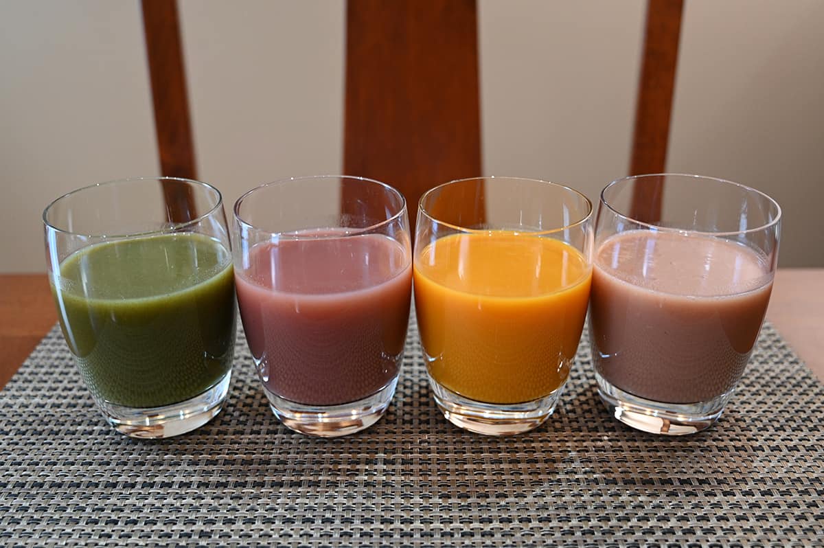 Image of four glasses with smoothie poured in it, from left to right a green smoothie, dark red smoothie, orange smoothie and pink smoothie.
