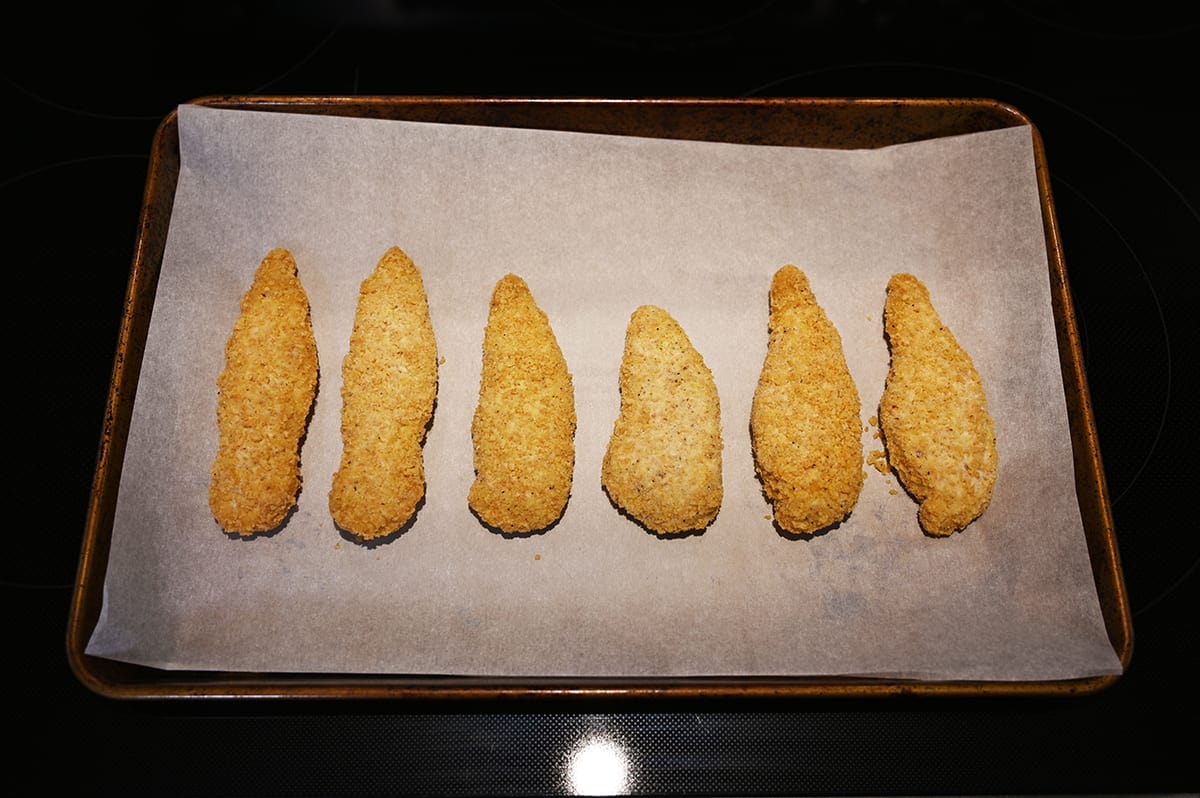 Image of six chicken strips on a baking sheet lined with parchment paper prior to baking them in the oven.