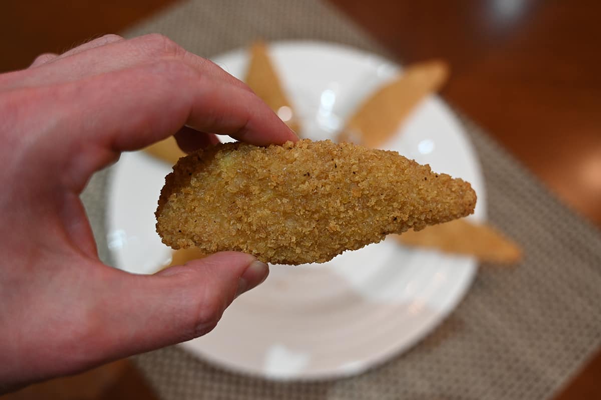 Closeup image of a hand holding one chicken strip in front of the camera with a plate of chicken strips in the background.
