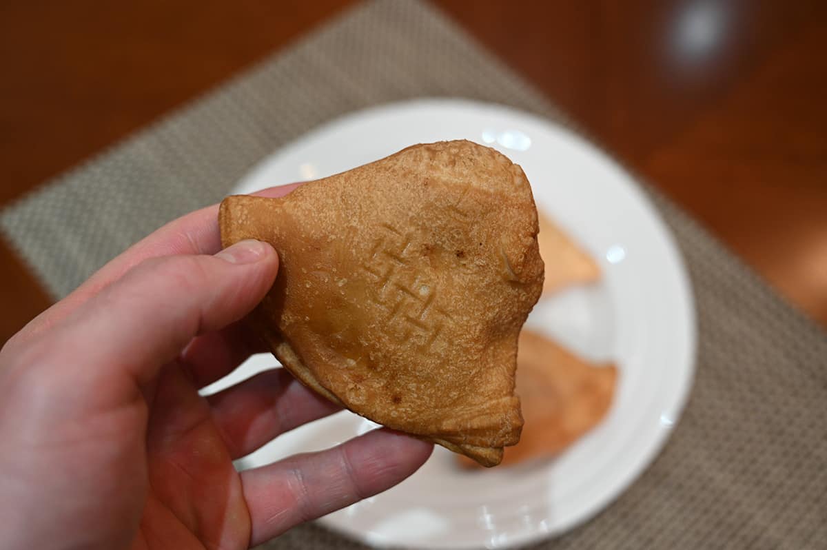 Image of a hand holding one samosa close to the camera with a plate of samosas in the background.
