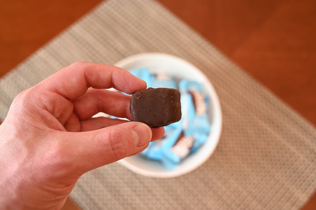 Closeup image of a hand holding one unwrapped coconut mini bar close to the camera with a bowl of bars in the baxkground.