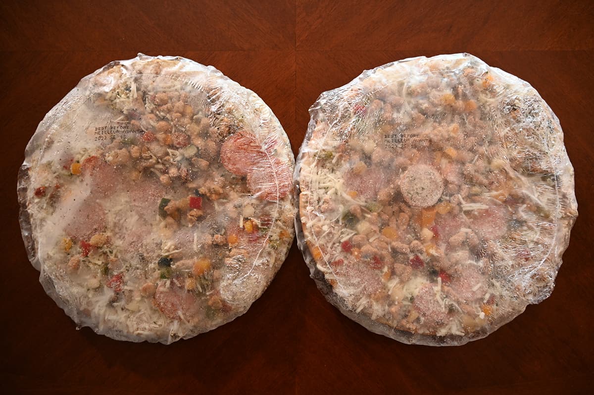 Top down image of two frozen pizzas individually packaged in plastic wrap sitting on a table.