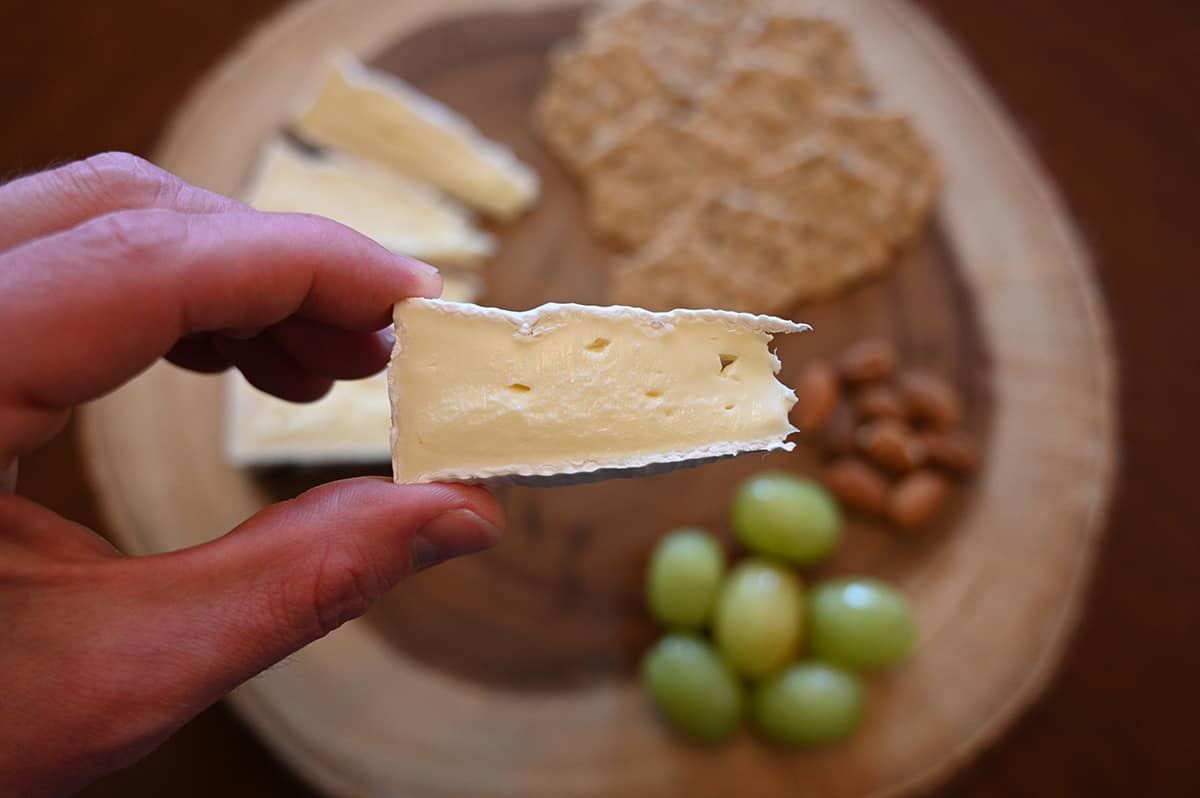 Closeup image of a hand holding one slice of Brie with a cheese board in the background of the image with cheese, crackers, nuts and grapes on it.