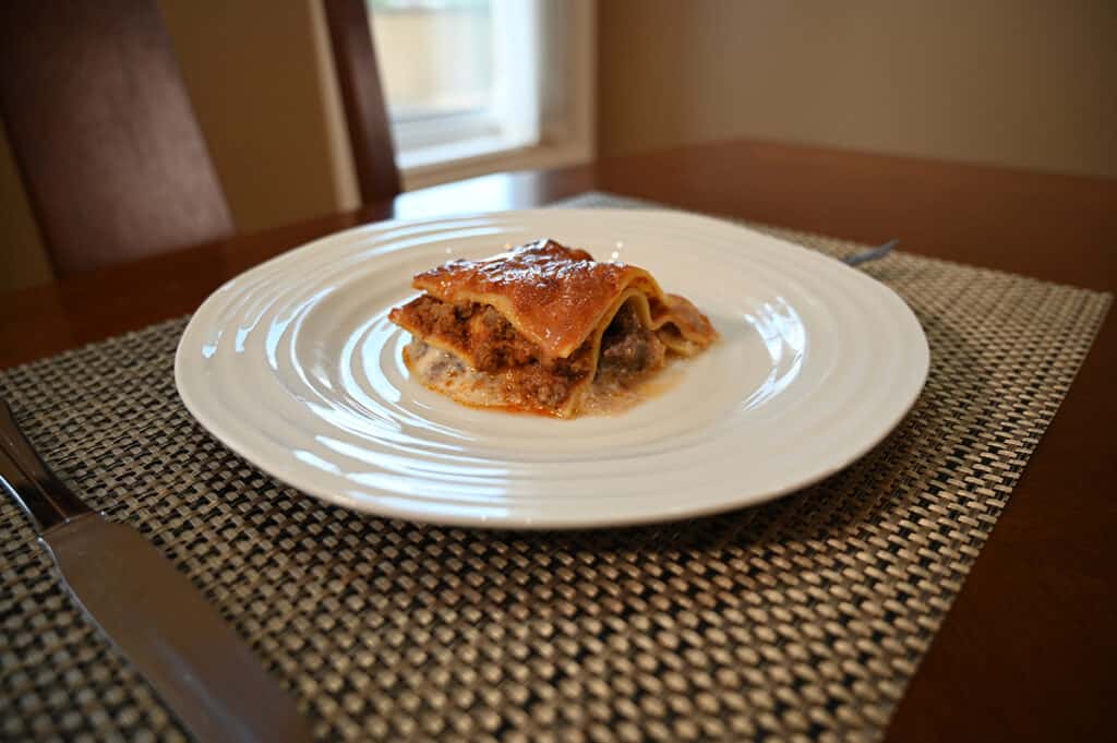 Side view image of a piece of cooked lasagna served on a white plate and sitting on a dining table.