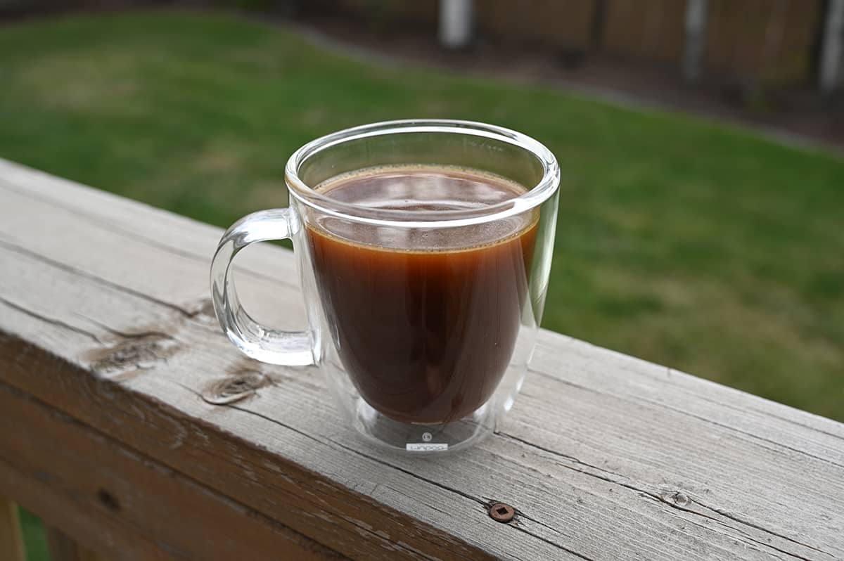 Image of a clear glass mug of coffee with cream sitting on a deck with grass in the background.