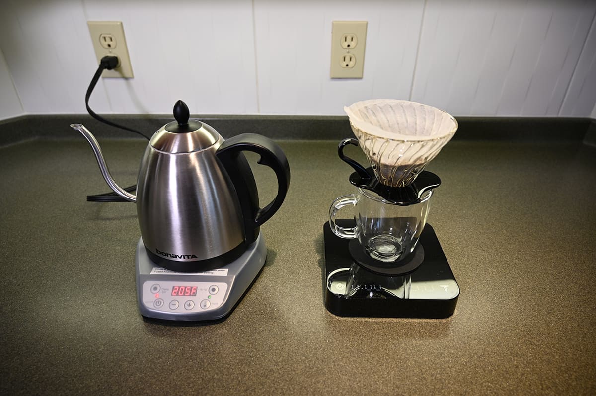 Far away image of coffee beans in a filter sitting over a glass mug on a kitchen scale with a kettle beside the mug on a kitchen counter.