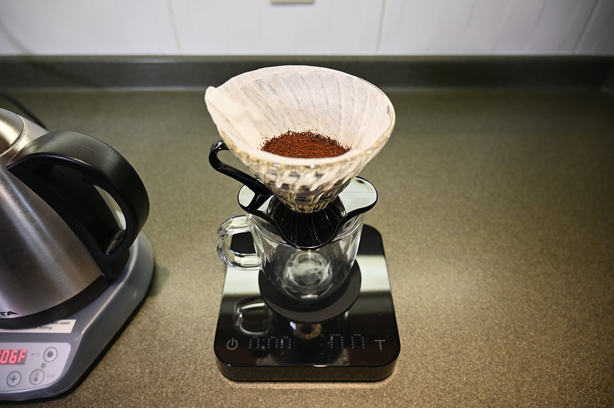 Image of ground coffee beans in a filter sitting over a glass mug on a kitchen scale with a kettle beside the mug. 