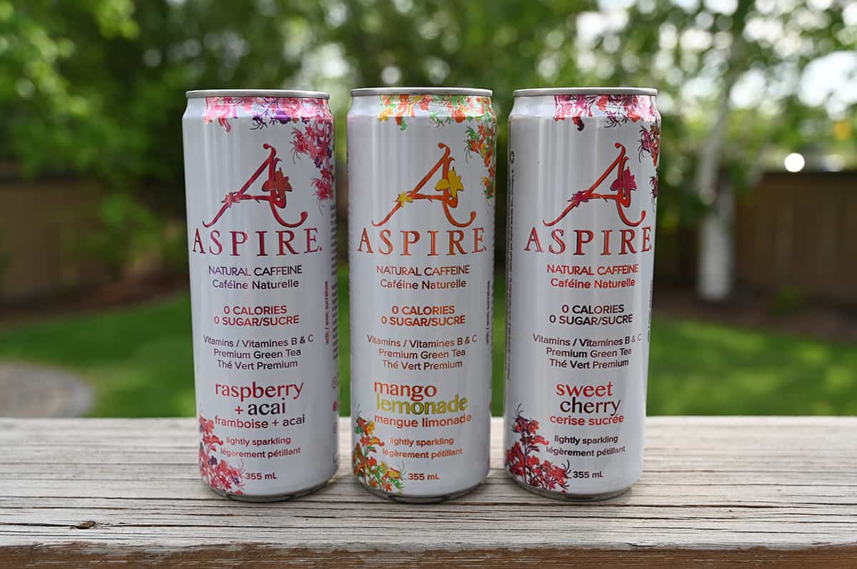 Image of three cans of Aspire Energy Drink sitting on a table outside. From left to right there is raspberry acai, mango lemonade and sweet cherry.
