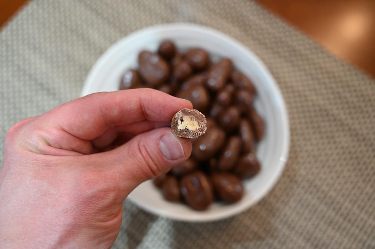 Image of one milk chocolate pecan with a bite taken out of it showing the inside, there is a bowl full of milk chocolate pecans in the background.