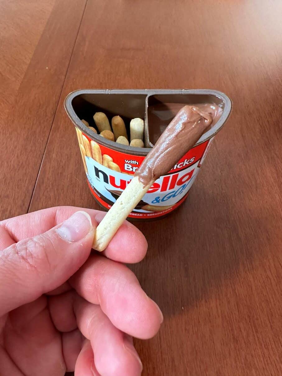 Closeup image of a hand holding one breadstick dipped in Nutella close to the camera with the package in the background.