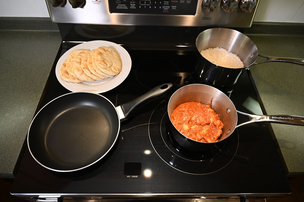 Top down image of a stove top with the vindaloo being heated in a pot as well as rice in a pot there is an empty frying pan and a plate of naan bread beside the frying pan.