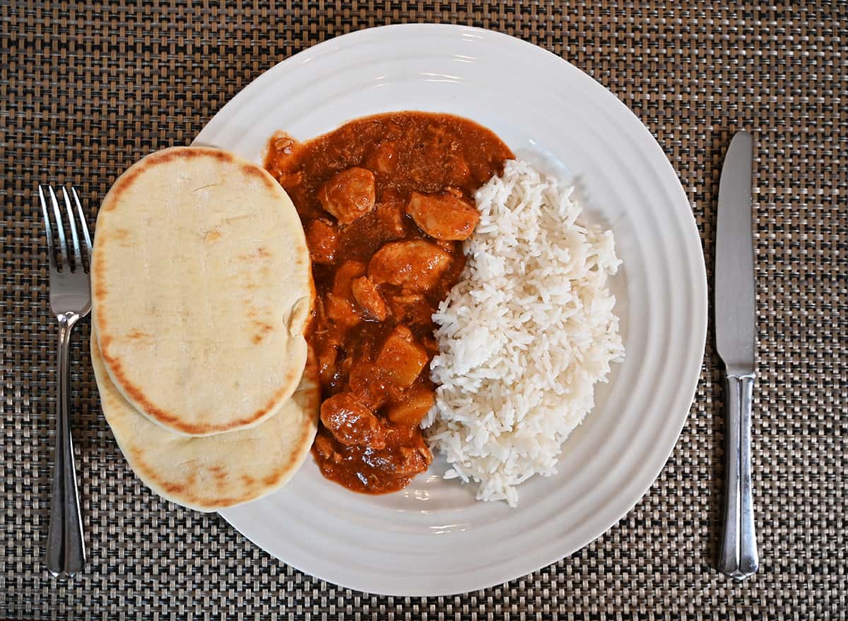 Top down image of a white plate with vindaloo, naan bread and rice on it. Beside the plate is a fork and knife.