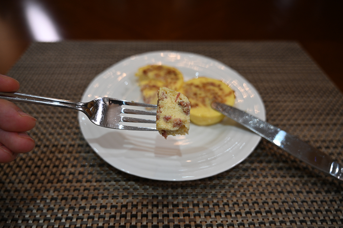 Side view closeup image of a fork full of cheese & uncured bacon egg bite there is a knife resting on the egg bite and there are two egg bites on a white plate.