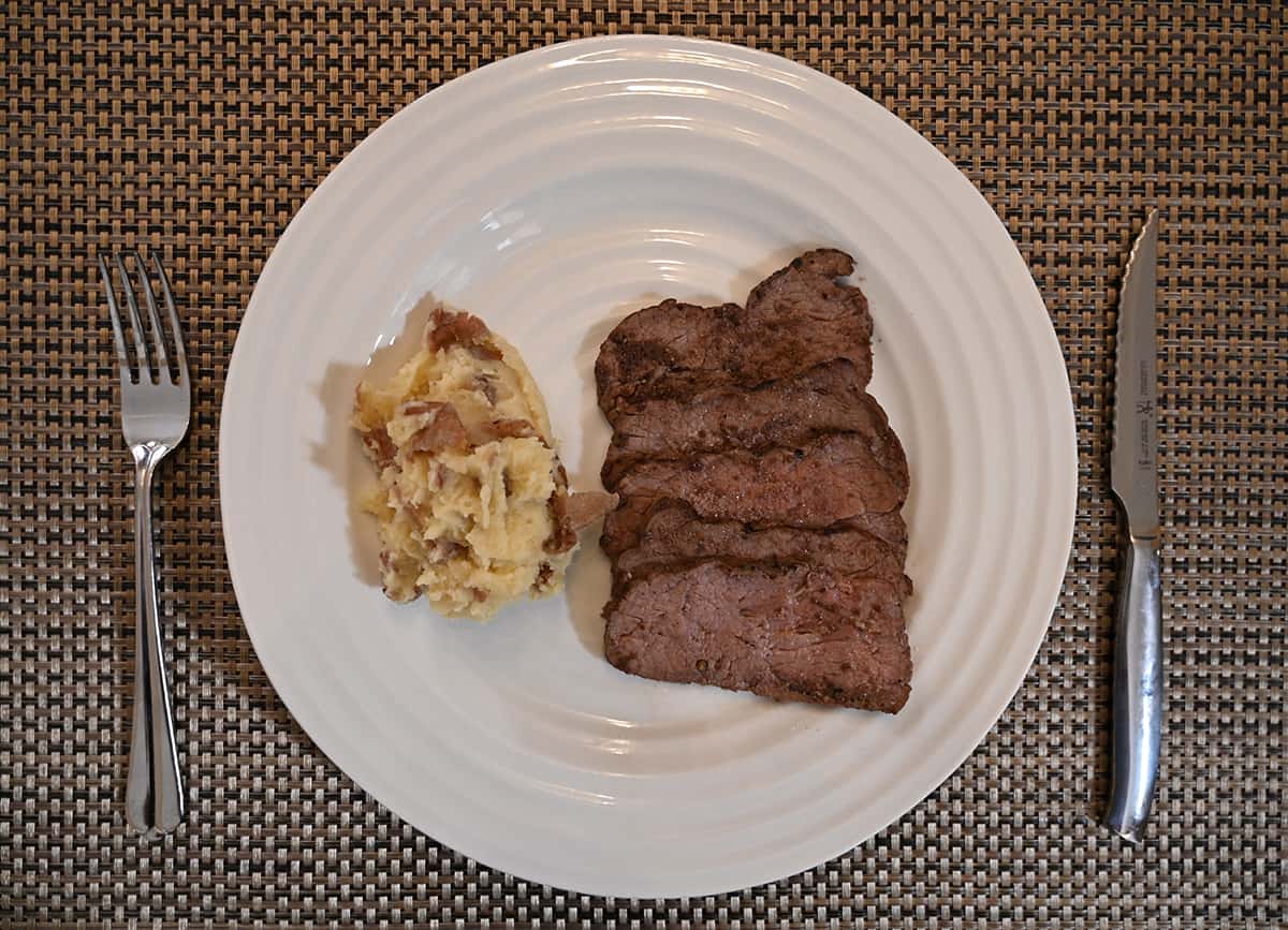 Top down image of a white plate with cooked sirloin on it beside a serving of mashed potatoes, there is a fork and knife beside the plate.