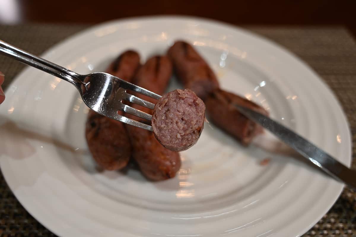 Image of cooked brats on a white plate. The brat is cut and a fork is facing the camera with a piece of brat on it so you can see the center of the brat.