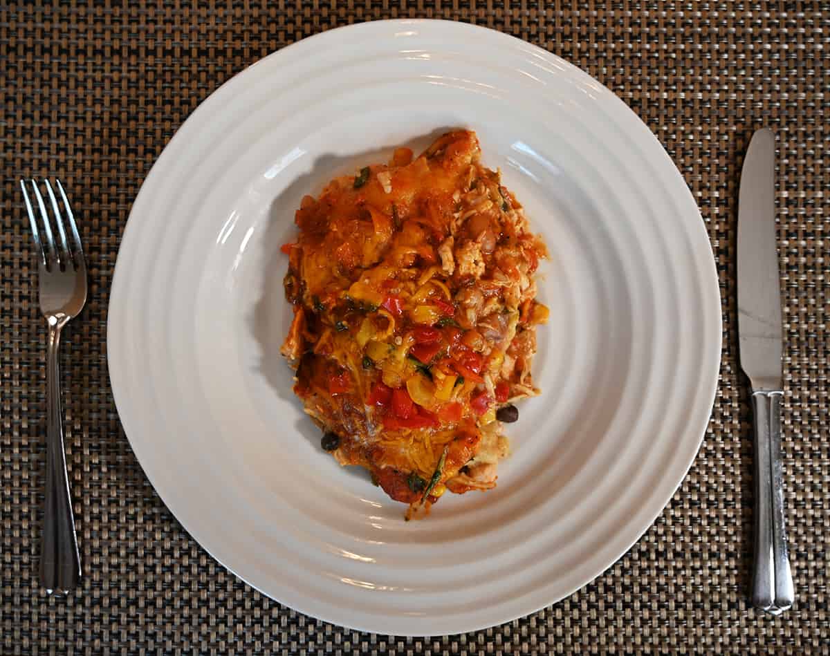 Top down image of a white plate with a piece of enchilada bake served on it. Beside the plate is a knife and a fork.