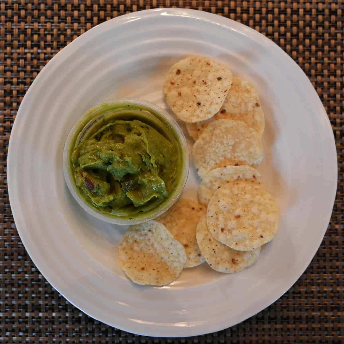Top down image of a white plate with tortilla chips on it and beside the chips is an open guacamole cup.