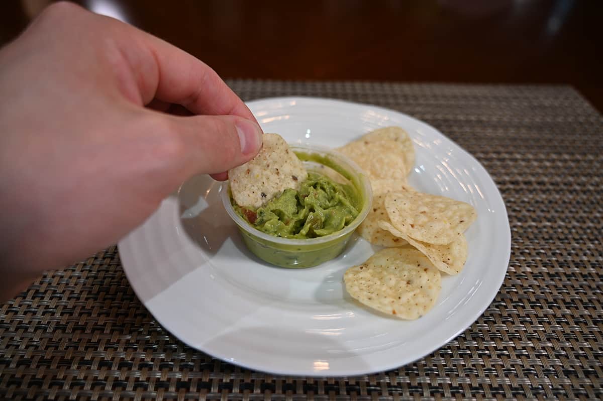 Image of a white plate with tortilla chips on it and beside the chips is a guacamole cup. There is a hand holding a chip and dipping it into the guacamole.