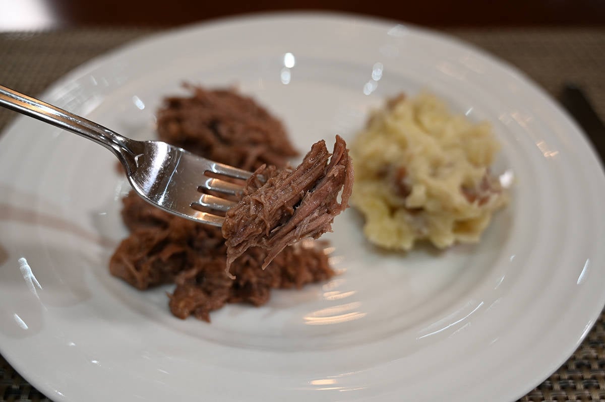 Side view closeup image of a fork with a bite of pot roast on it, in the background is a plate with pot roast and mashed potatoes on it.