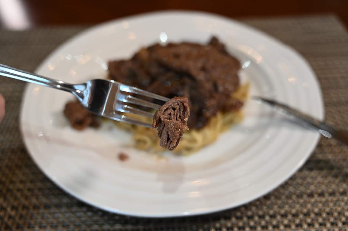 Side view image of a piece of beef bulgogi on a fork with a plate of bulgogi and noodles in the background.