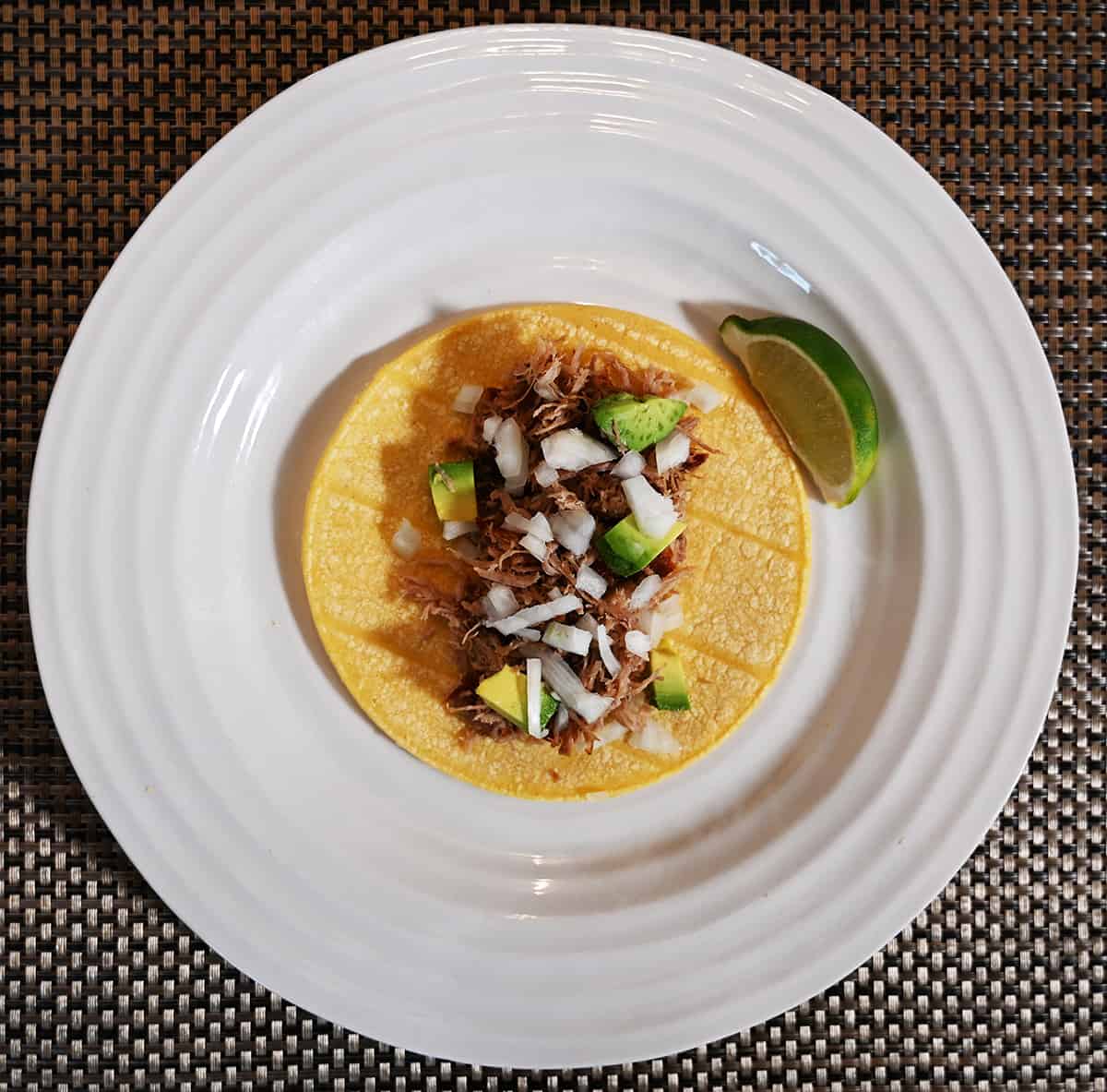 Top down image of a prepared carnita taco open with avocado and onions on it with a slice of lime beside it.