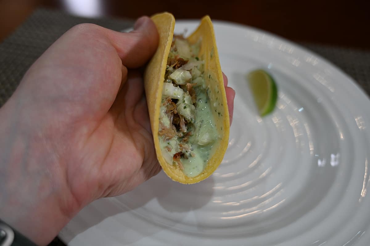 Closeup image of a hand holding a pork carnita close to the camera with cilantro lime crema drizzled on the pork. There is a white plate with a wedge of lime in the background.