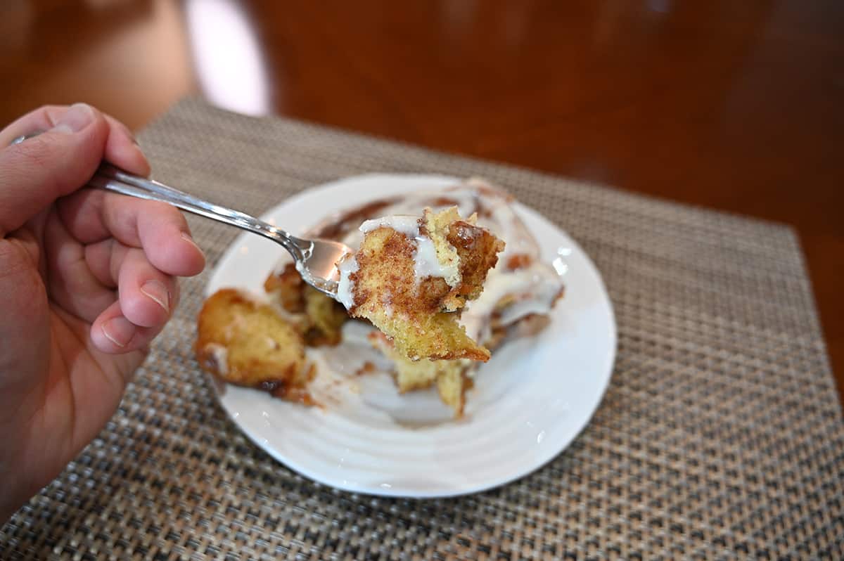 Closeup side view image of a fork with a piece of cinnamon roll on it, in the background is a white plate with a cinnamon roll on it.
