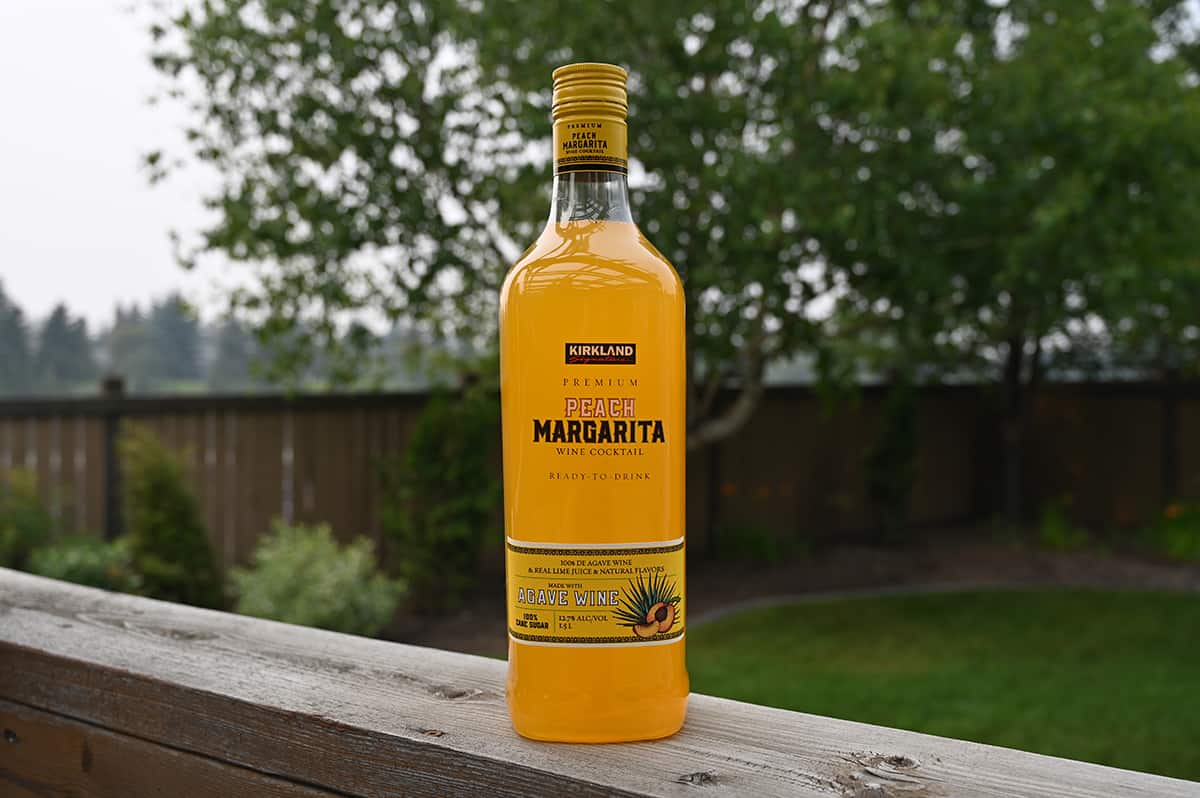 Image of the Kirkland Signature Premium Peach Margarita Wine Cocktail standing on a deck with trees, grass and a fence in the background.