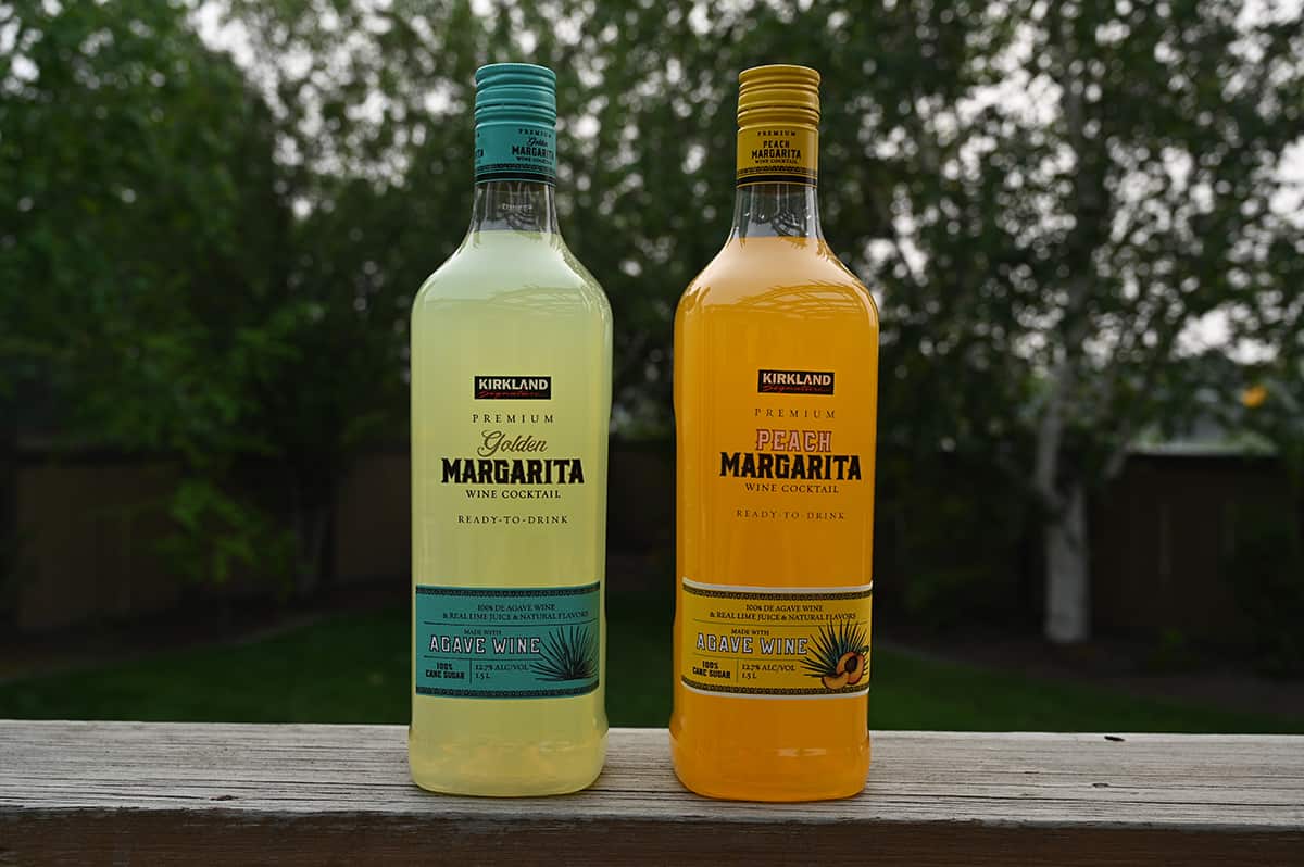 Image of the Costco Kirkland Signature Margarita Wine Cocktails in Golden Margarita and Peach standing side by side on a deck there are trees in the background.