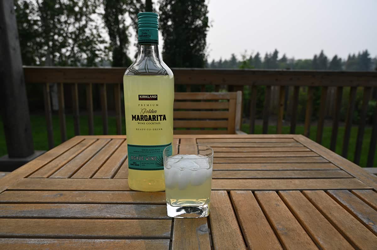 Image of the Kirkland Signature Premium Golden Margarita Wine Cocktail bottle standing on a deck  beside a glass of poured cocktail with trees, grass and a fence in the background.