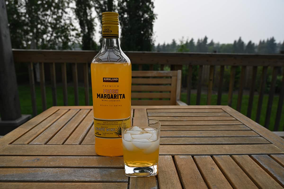 Image of the Kirkland Signature Premium Peach Margarita Wine Cocktail bottle standing on a deck  beside a glass of poured cocktail with trees, grass and a fence in the background.