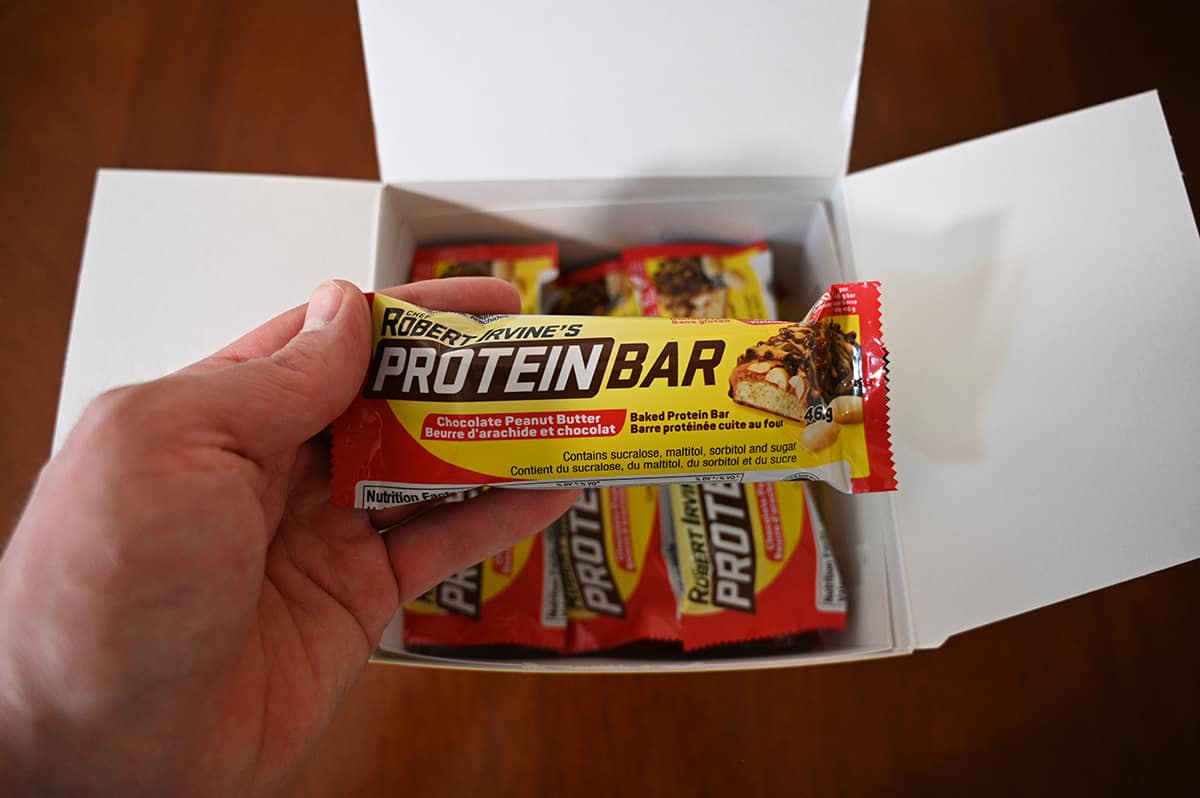 Image of a hand holding one individually packaged protein bar over top an open box of protein bars.