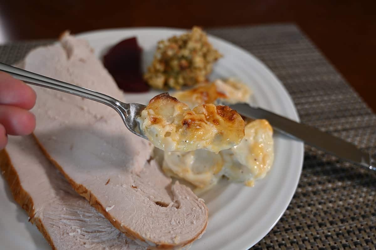 Closeup image of a fork with a mouthful of scalloped potatoes served on it. In the background of the image is turkey, cranberry sauce and stuffing.