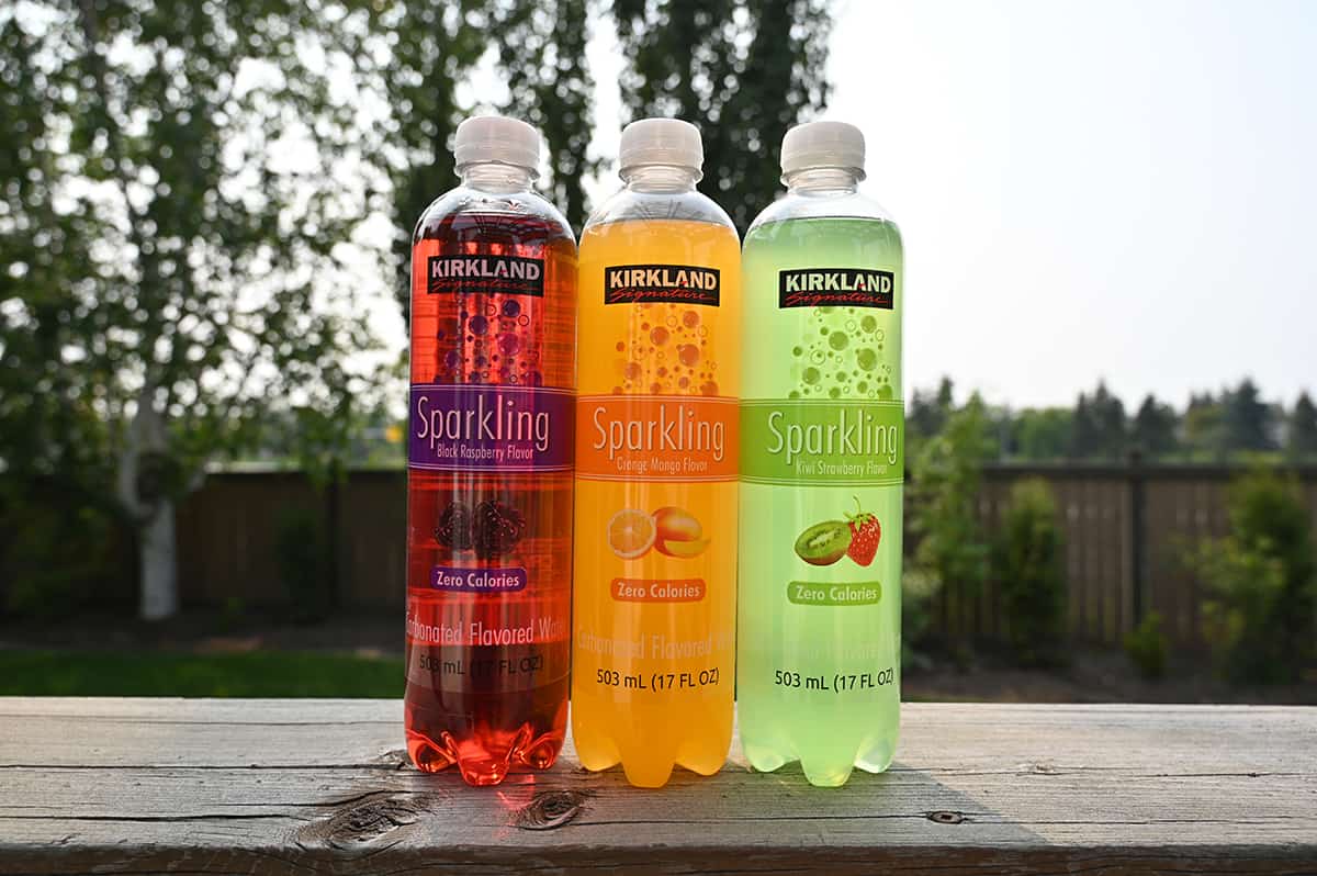 Image of three full bottles of Kirkland Signature Sparkling Water sitting on a deck,there is black raspberry, mango orange and kiwi strawberry.