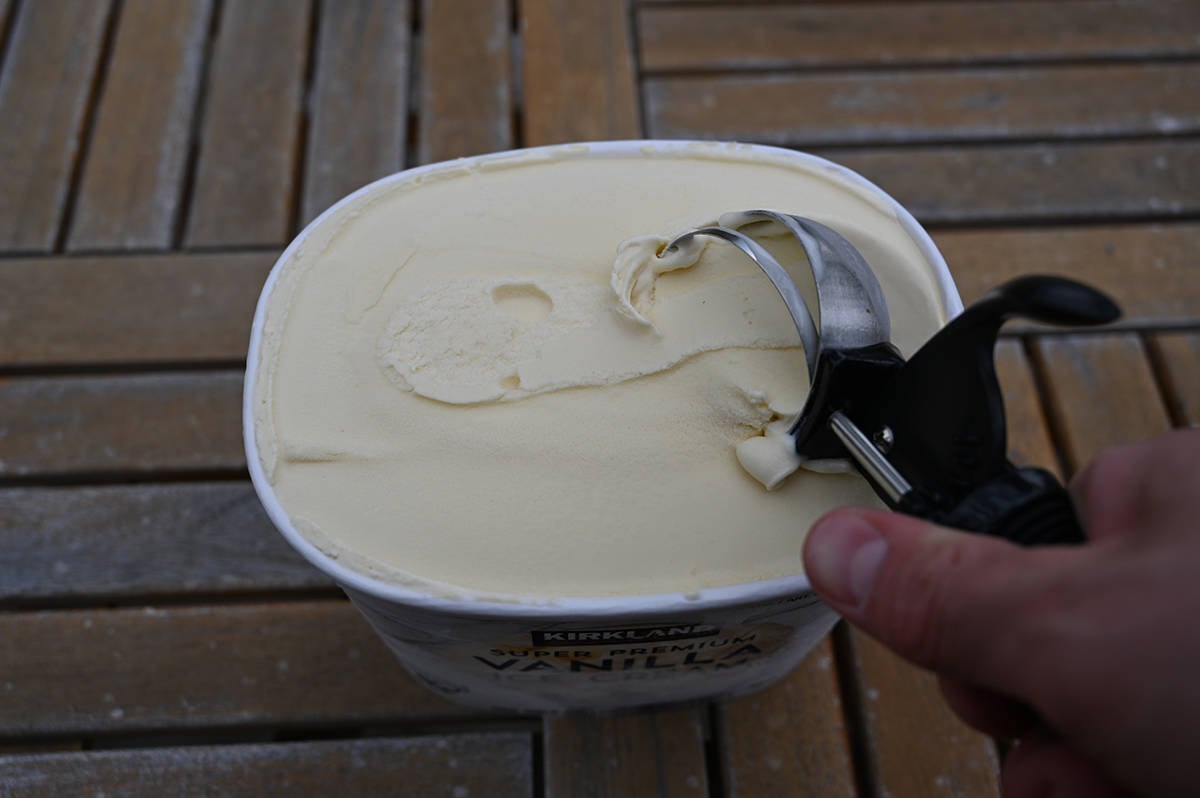 Image of a tub of vanilla ice cream opened with a hand holding a scoop in the ice cream.