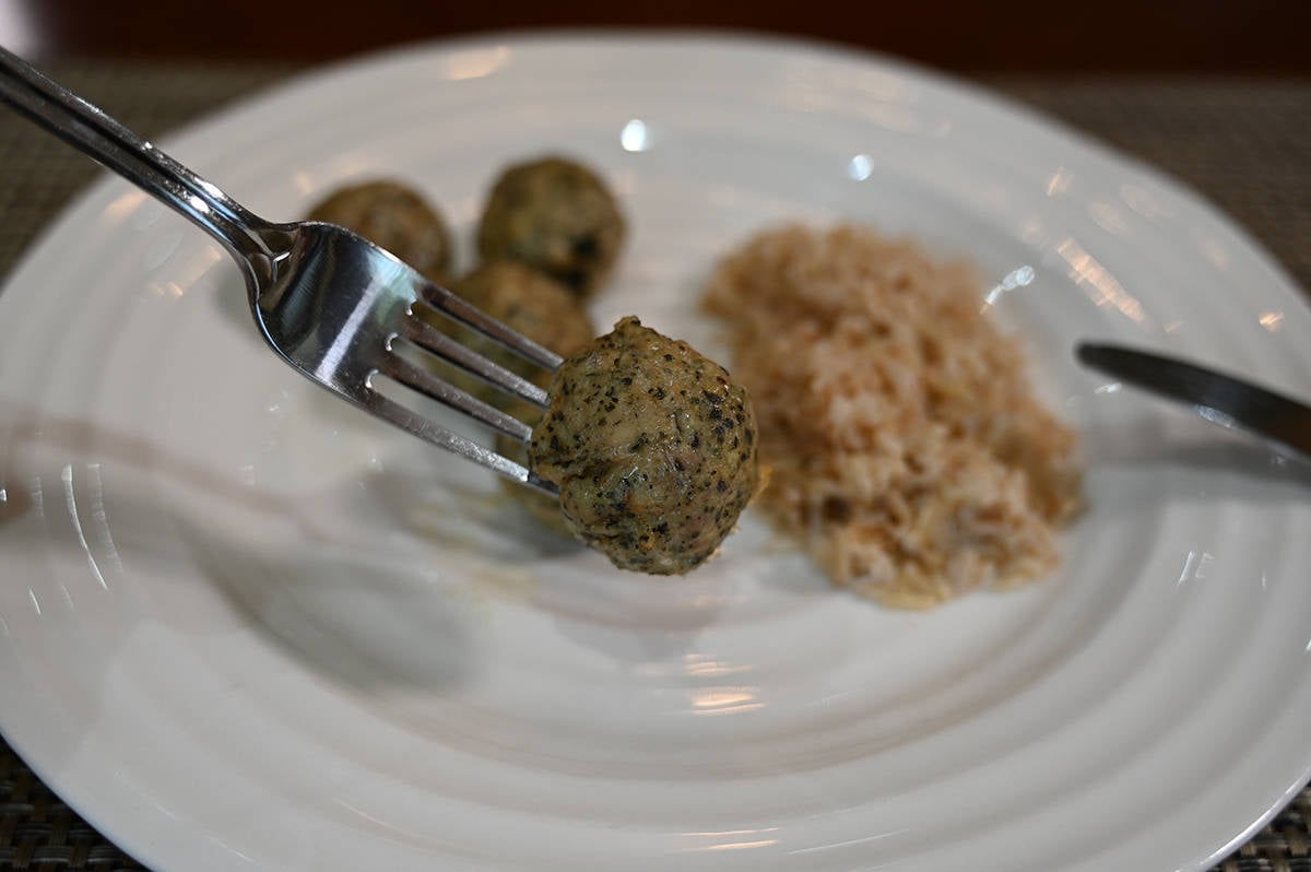 Image of a fork with a whole meatball on it so you can see the center of the meatball, in the background is a plate with more meatballs and rice on it.