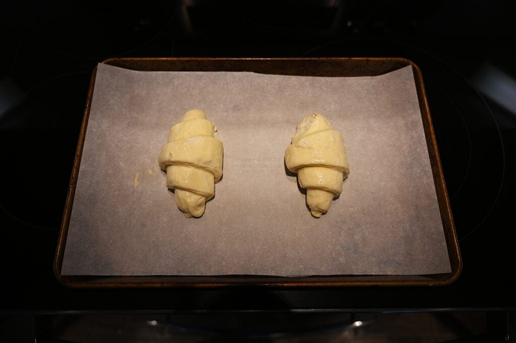 Top down image of two unbaked croissants sitting on a parchment lined baking tray on top of an oven.
