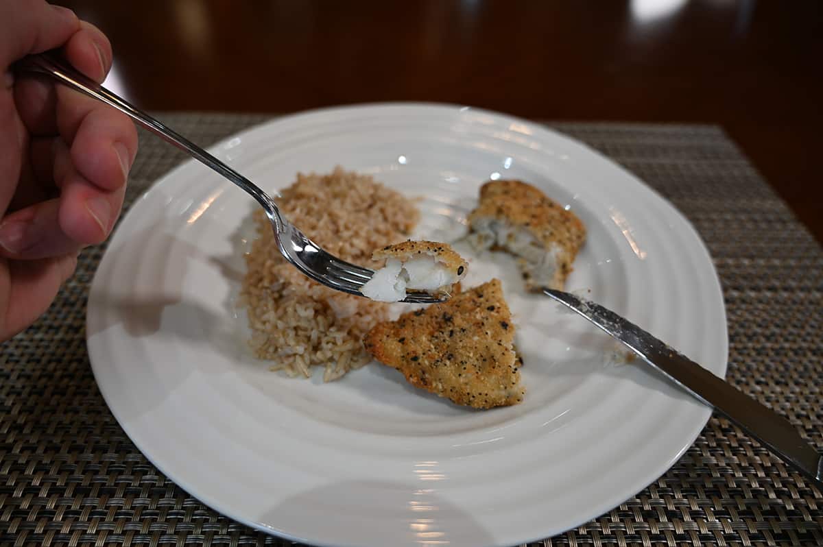 Image of a plate of baked cod beside a bed of rice, there is a fork hovering above the plate with a bite of cod on the fork.