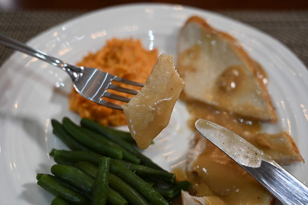 Closeup image of a fork with a piece of turkey with gravy on it sitting on the fork. In the background is a plate with green beans, mashed potatoes and turkey.