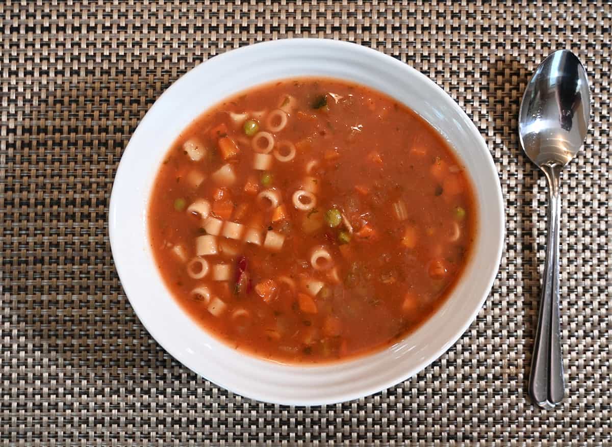 Top down closeup image of one large bowl of prepared vegetable minestrone soup in a white bowl with a spoon beside it.