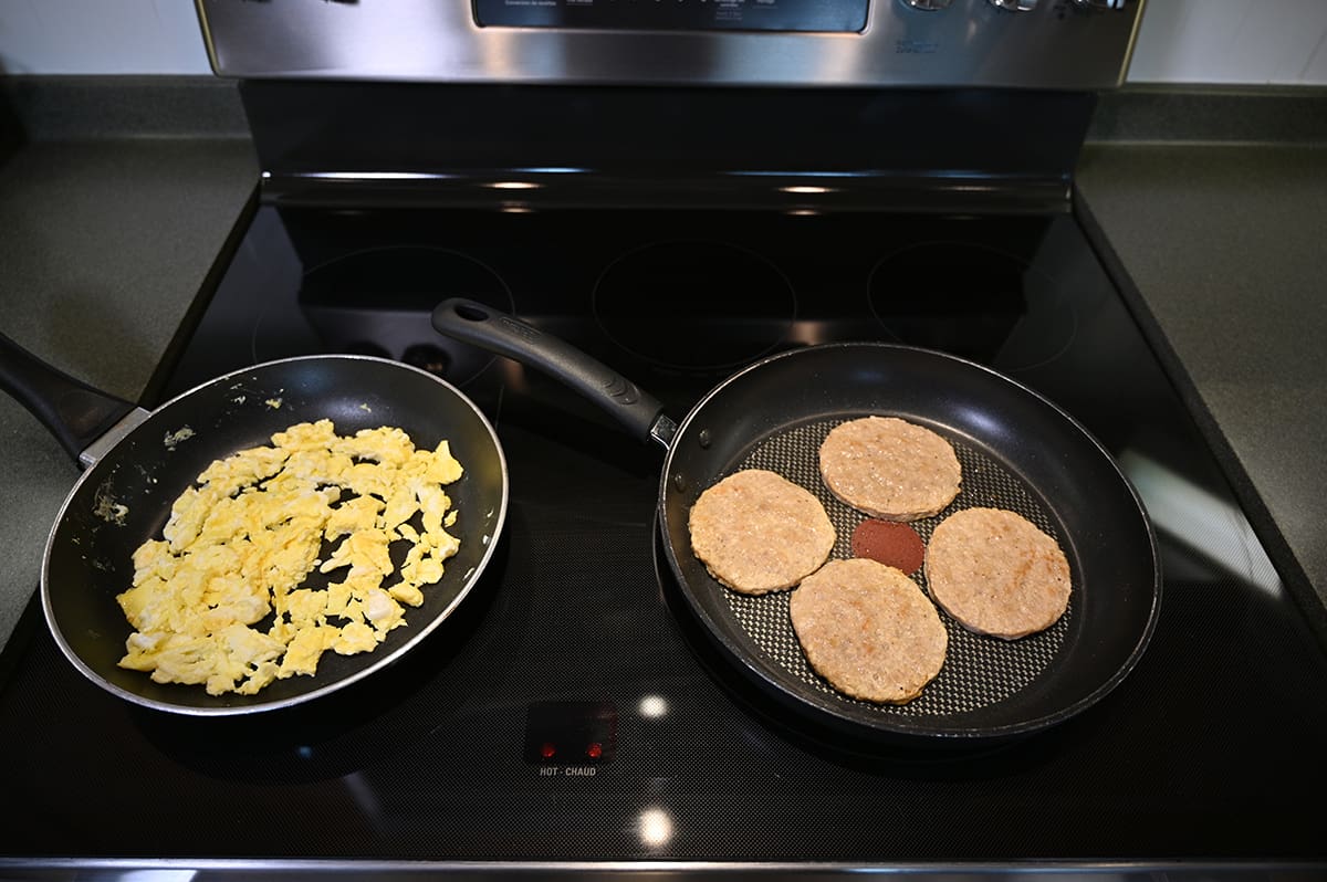 Top down image of a pan of scrambled eggs cooking beside a pan of four sausage rounds cooking on a stovetop.