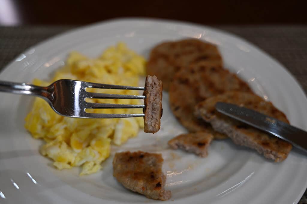 Closeup side view image of a fork with one bite of sausage round on it so you can see the center of the sausage. In the background is eggs and sausage.