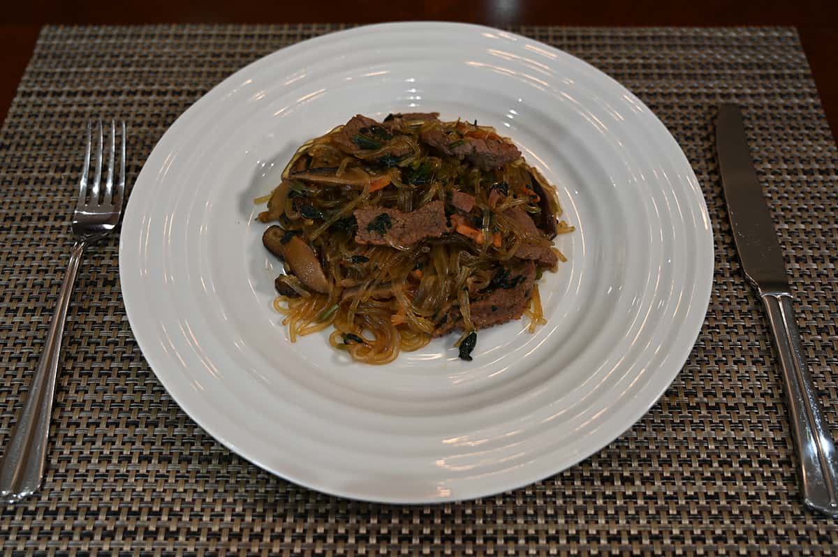 Sideview top down image of the japchae served on a white plate with a fork and knife beside the plate.