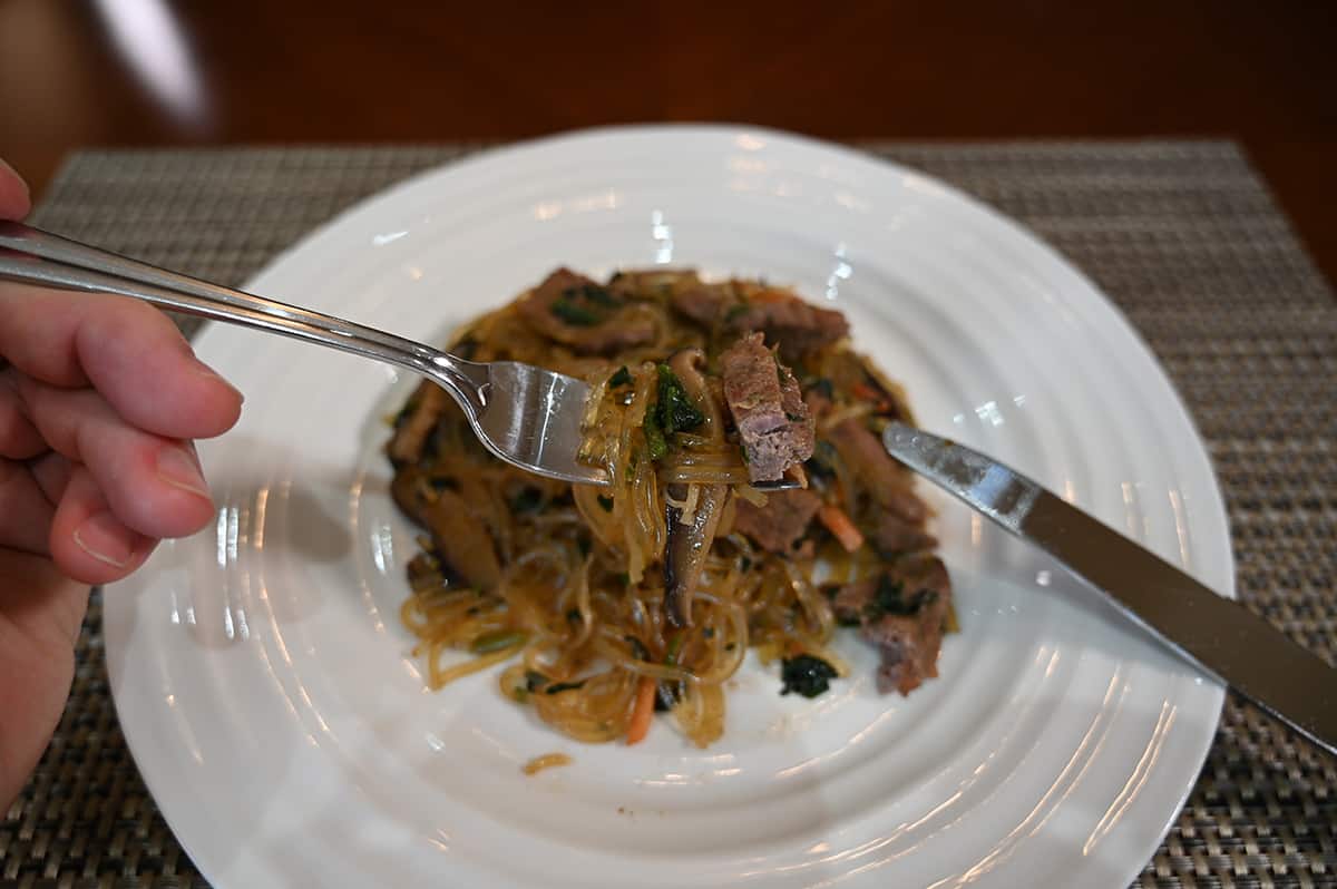 Top down closeup image of a hand holding a fork with Japchae on it close to the camera so you can see the beef and noodles.