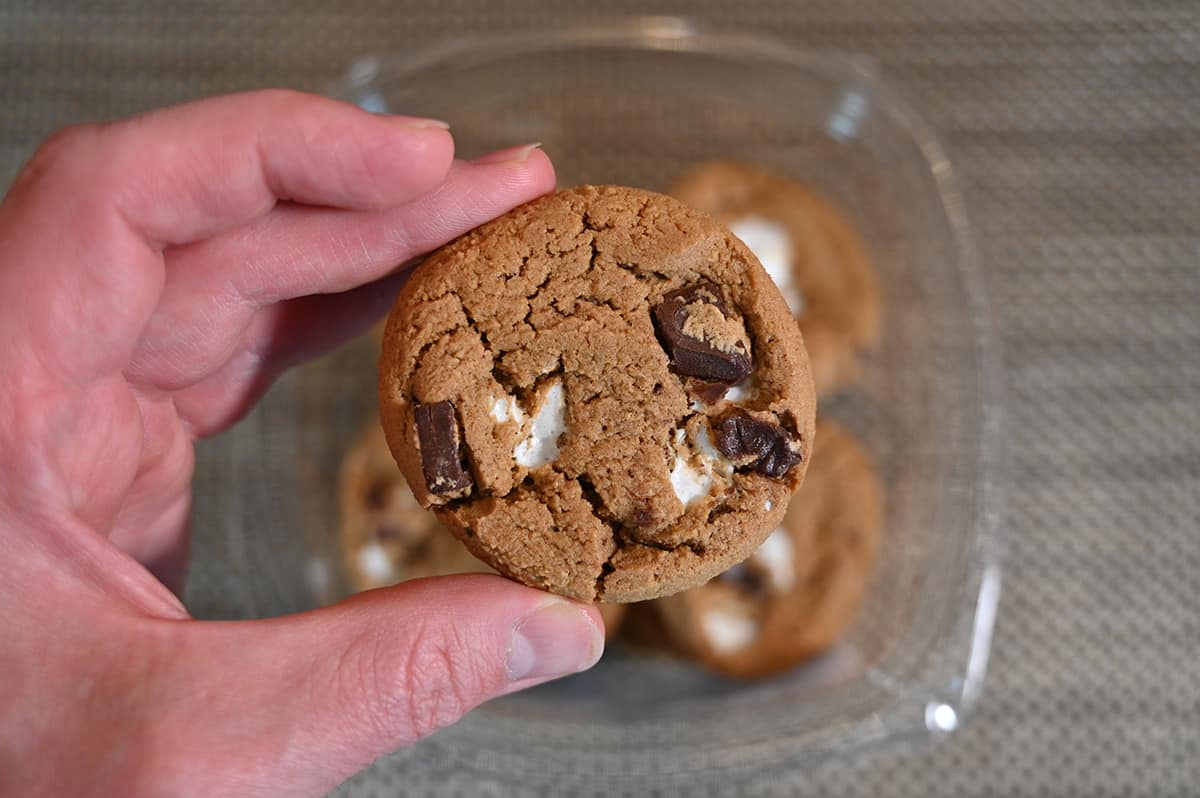 Closeup image of a hand holding one s'more cookie close to the camera with an open container of cookies in the background.