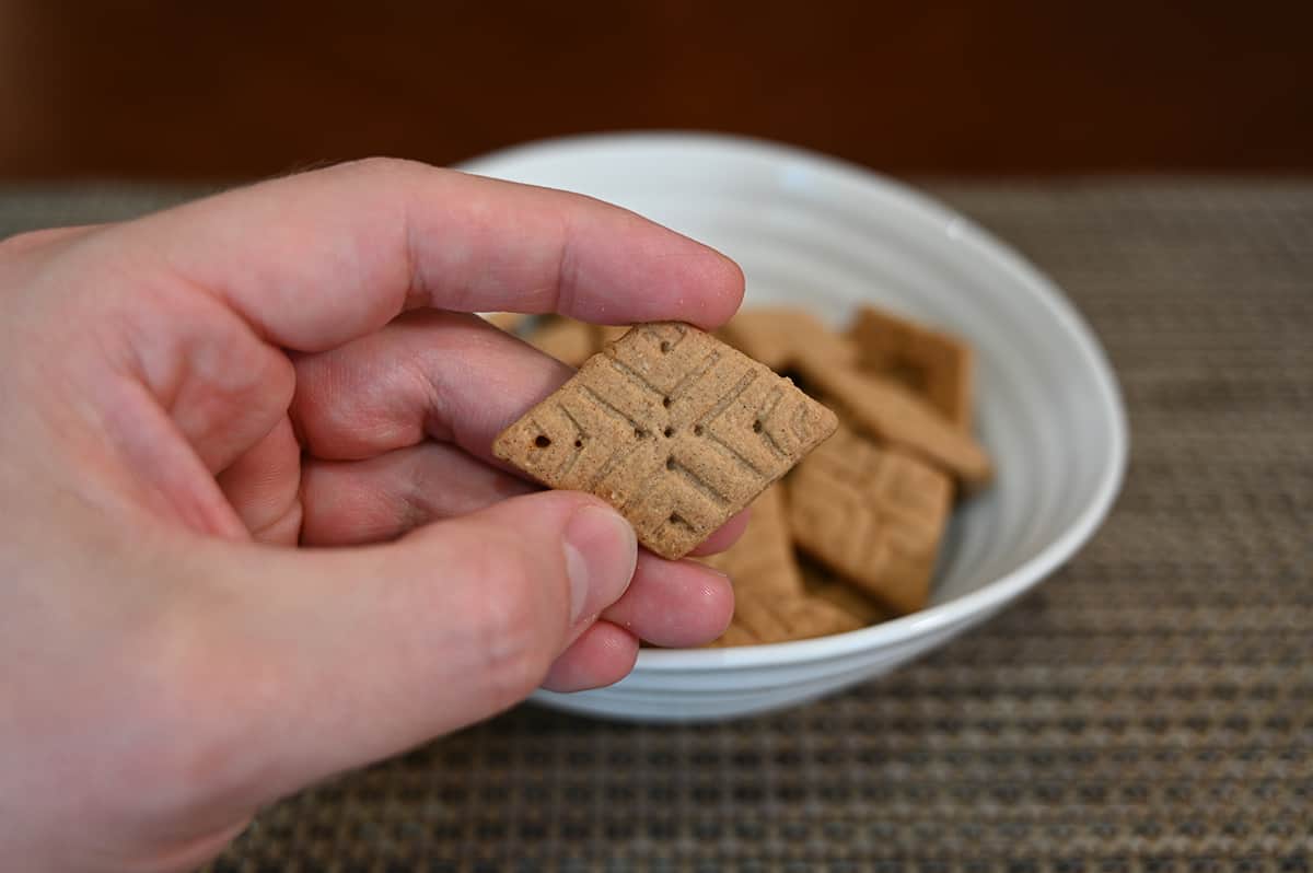 Closeup image of a hand holding one sweet thin close to the camera with a bowl of Sweet Thins in the background.