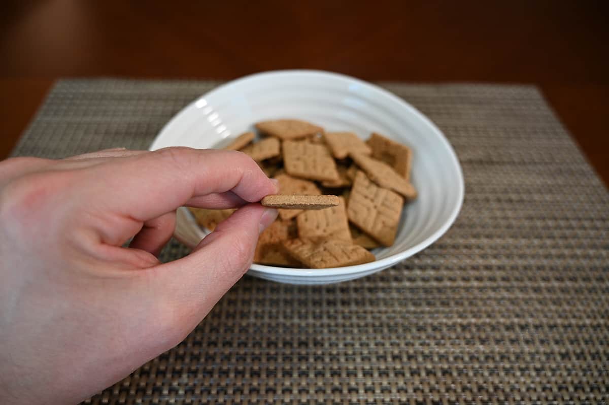 Closeup image of a hand holding one Sweet Thin angled on its side so you can see the width, there is a bowl of Sweet Thins in the background.