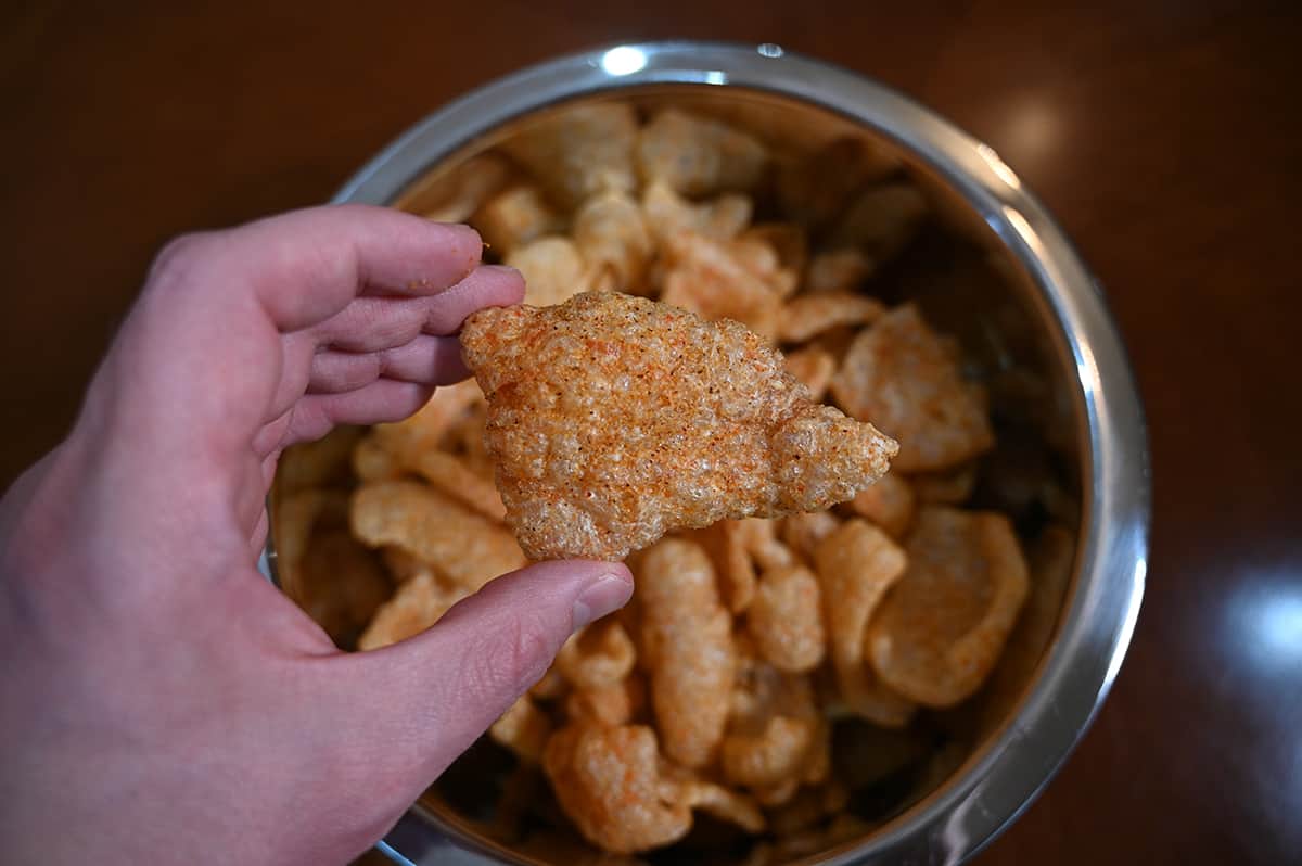 Top down image of a hand holding one pork rind hovering over a large metal bowl full of pork rinds.