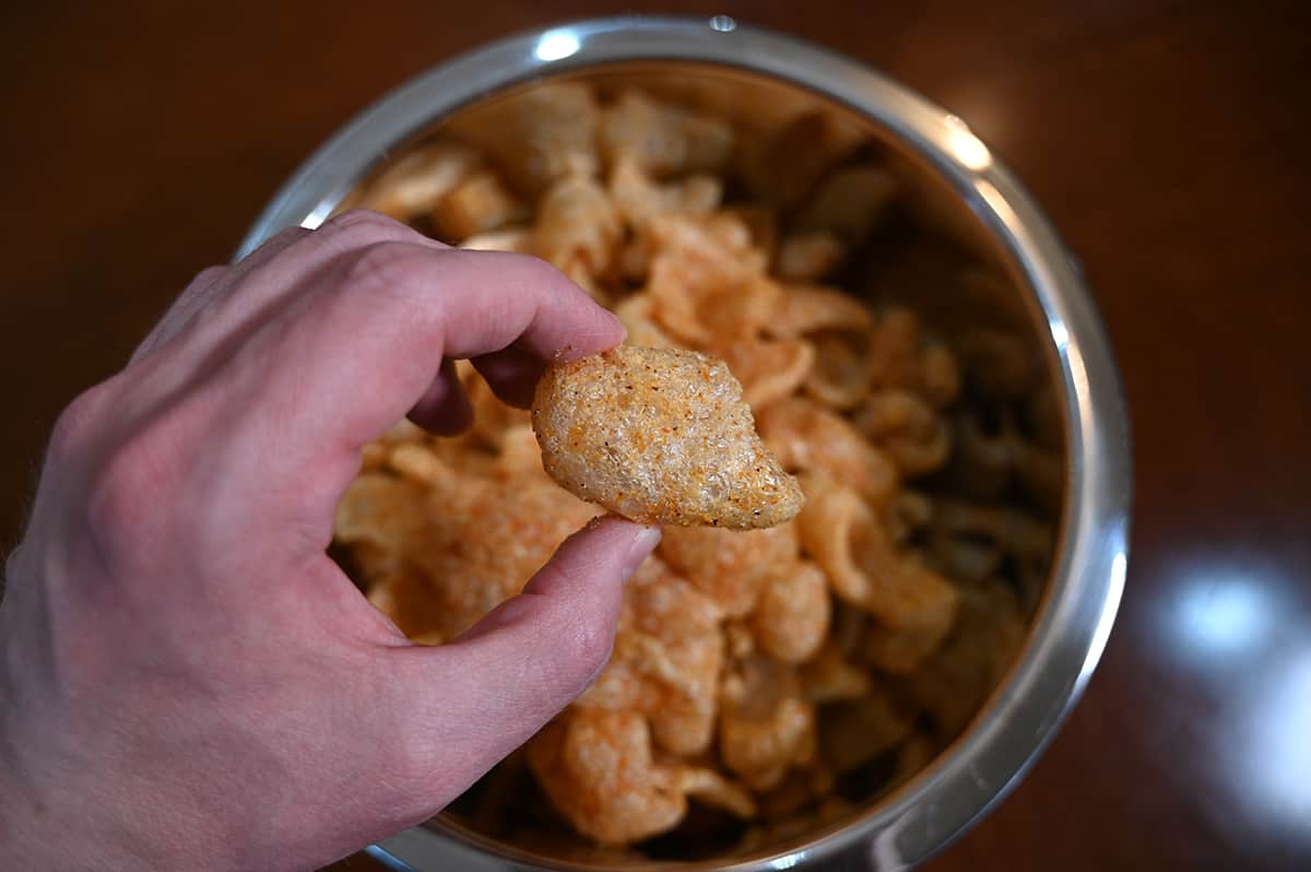 Top down image of a hand holding one pork rind hovering over a large metal bowl full of pork rinds.