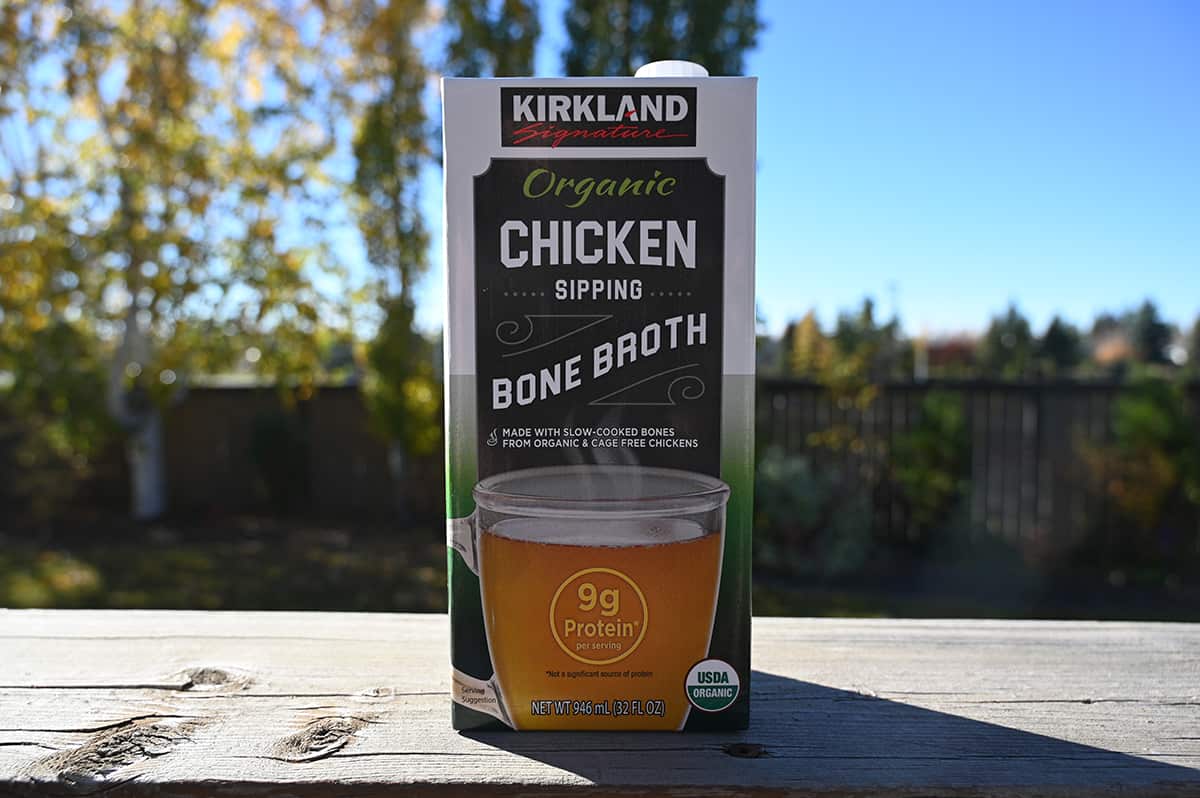 Image of one single carton of bone broth sitting on a deck outside with trees and a fence in the background.